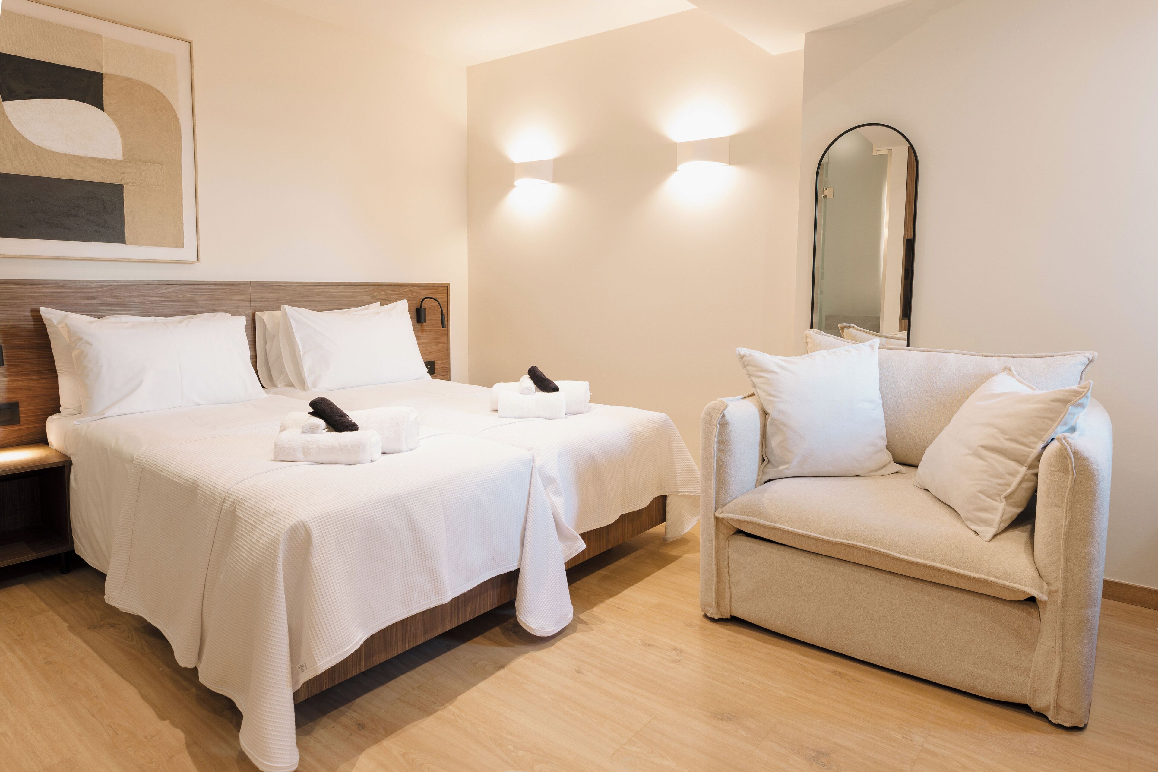 A hotel room with a bed, a chair, a bedside table, a mirror, and a wall-mounted light fixture.