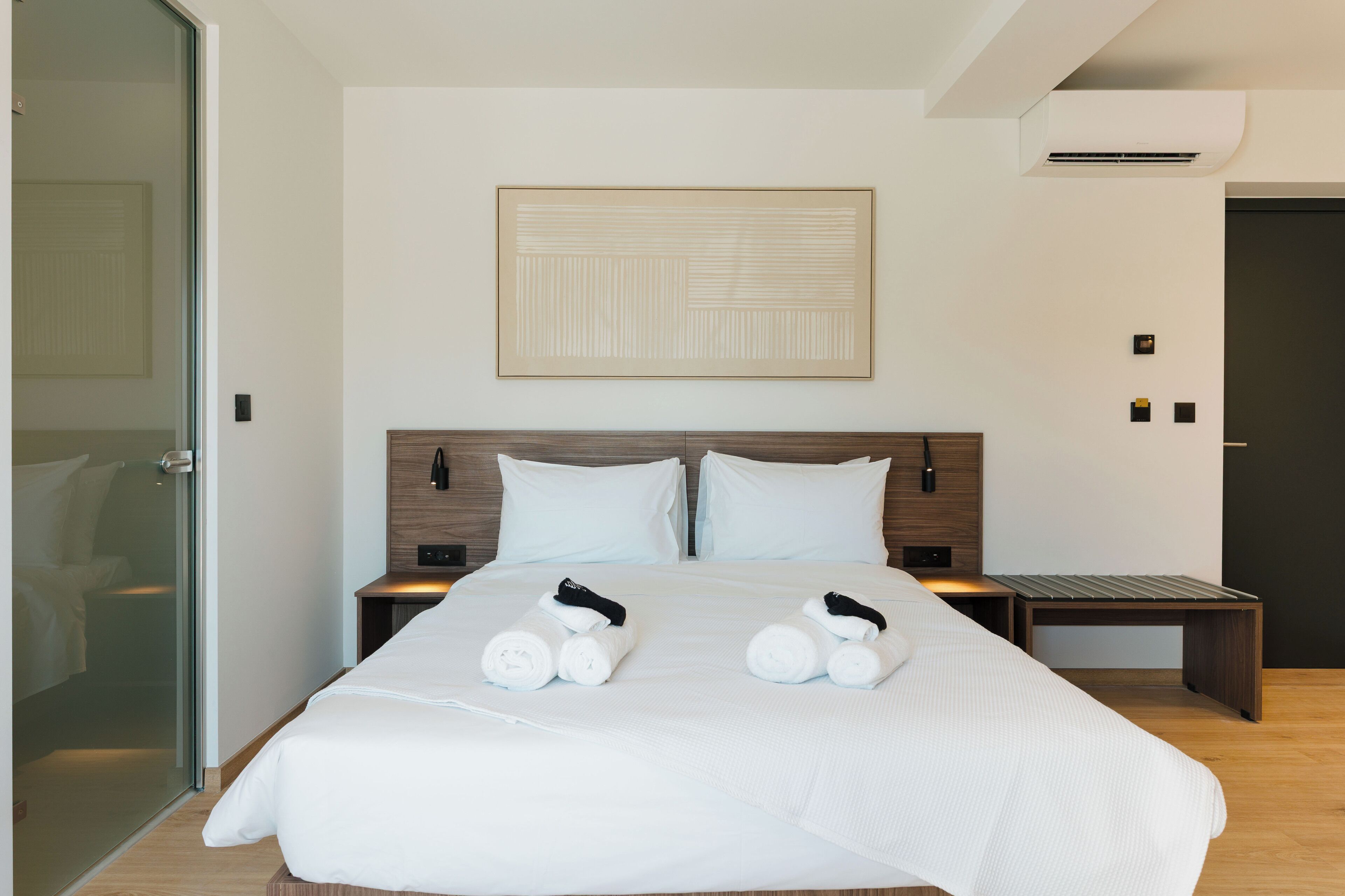 A modern hotel room with a large bed, two bedside tables, a wall-mounted artwork, and a glass shower door.