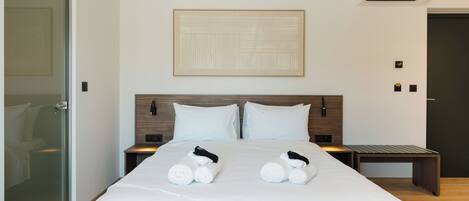 A modern hotel room with a large bed, two bedside tables, a wall-mounted artwork, and a glass shower door.
