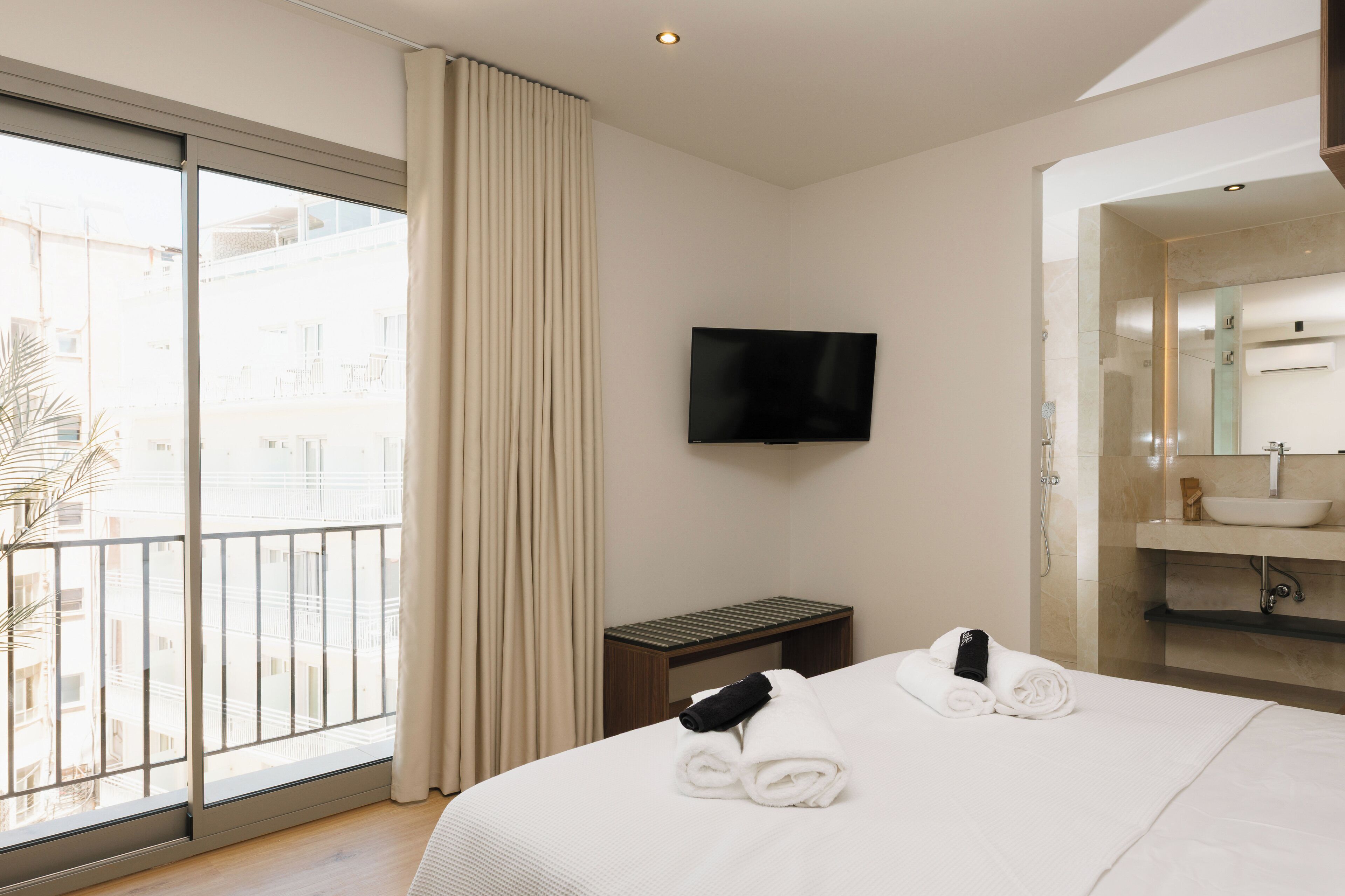 Une chambre d’hôtel dotée d’une grande fenêtre, d’un téléviseur à écran plat, d’un lit avec des serviettes soigneusement pliées, d’une petite table et d’une salle de bain avec un lavabo et un miroir.