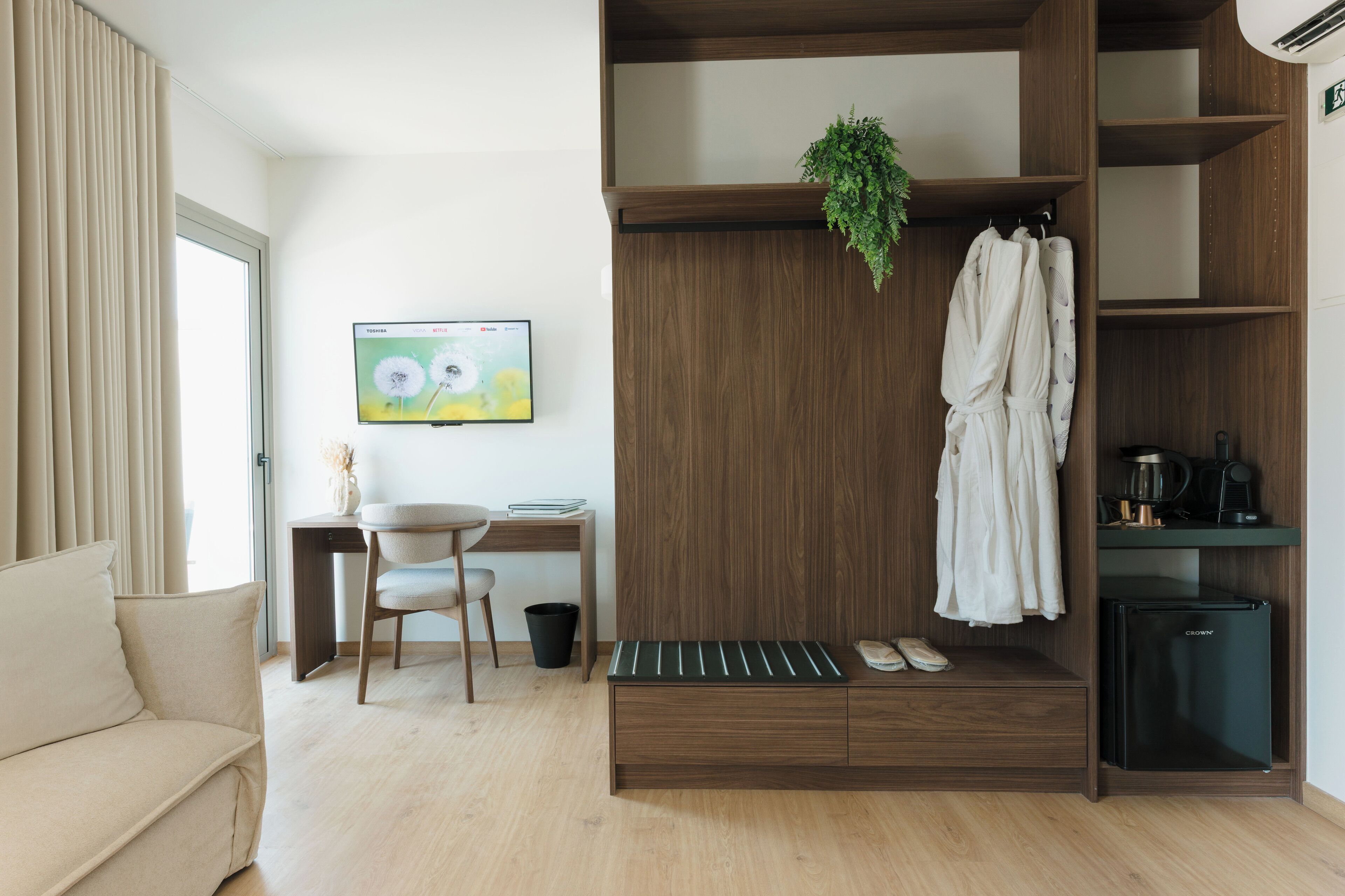 A modern hotel room with a wooden wardrobe, a small desk with a chair, a TV mounted on the wall, and a compact kitchenette.