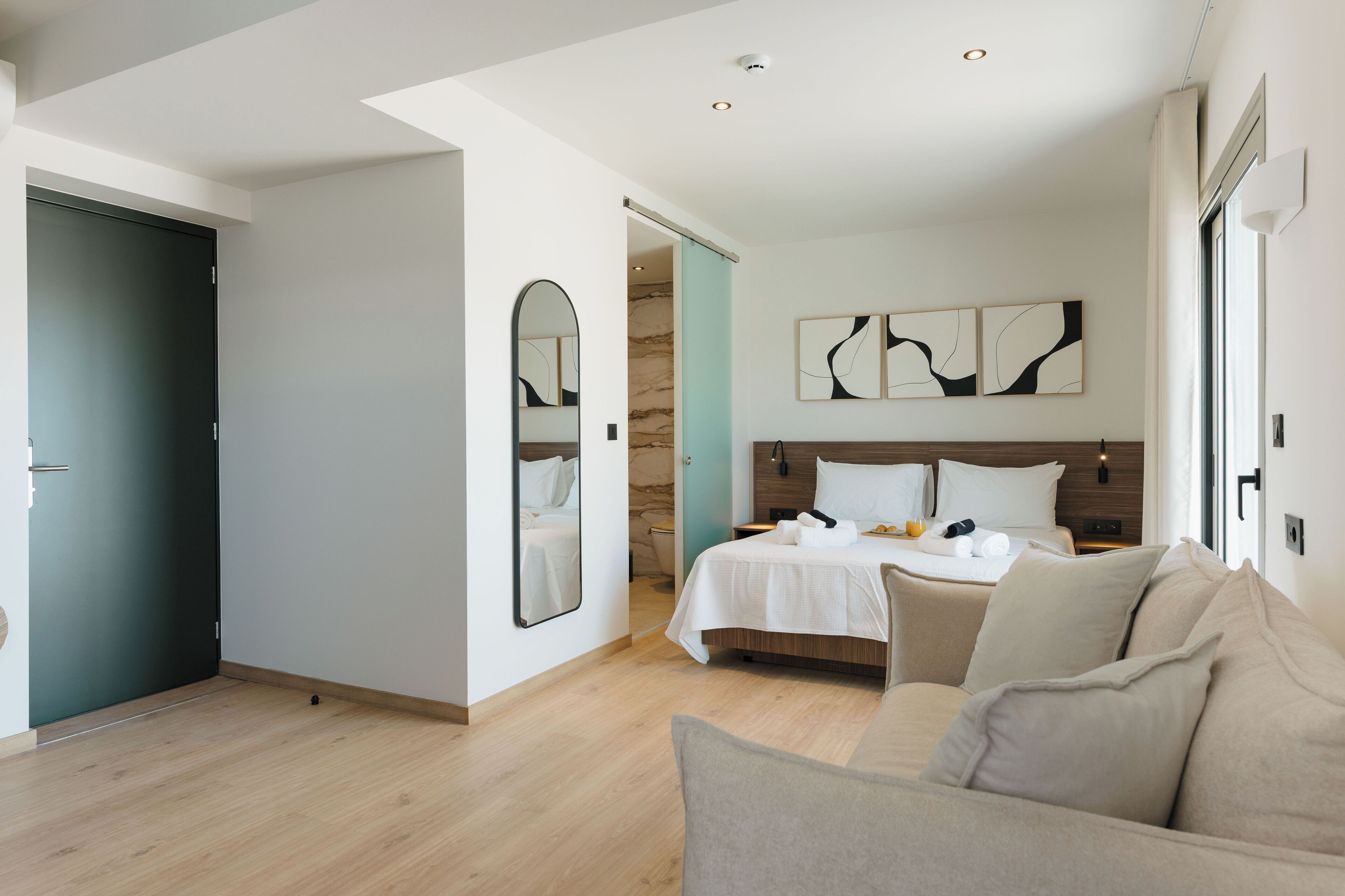 A modern hotel room with a bed, a sofa, a mirror, and abstract wall art.