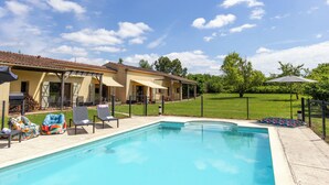 Piscina com espreguiçadeiras e guarda-sóis, cercada por um quintal e uma casa com área de estar externa.