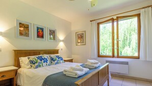 Um quarto com cama de madeira, roupa de cama branca, um travesseiro floral azul, uma mesa de cabeceira e cortinas na janela. Luzes fixadas na parede.