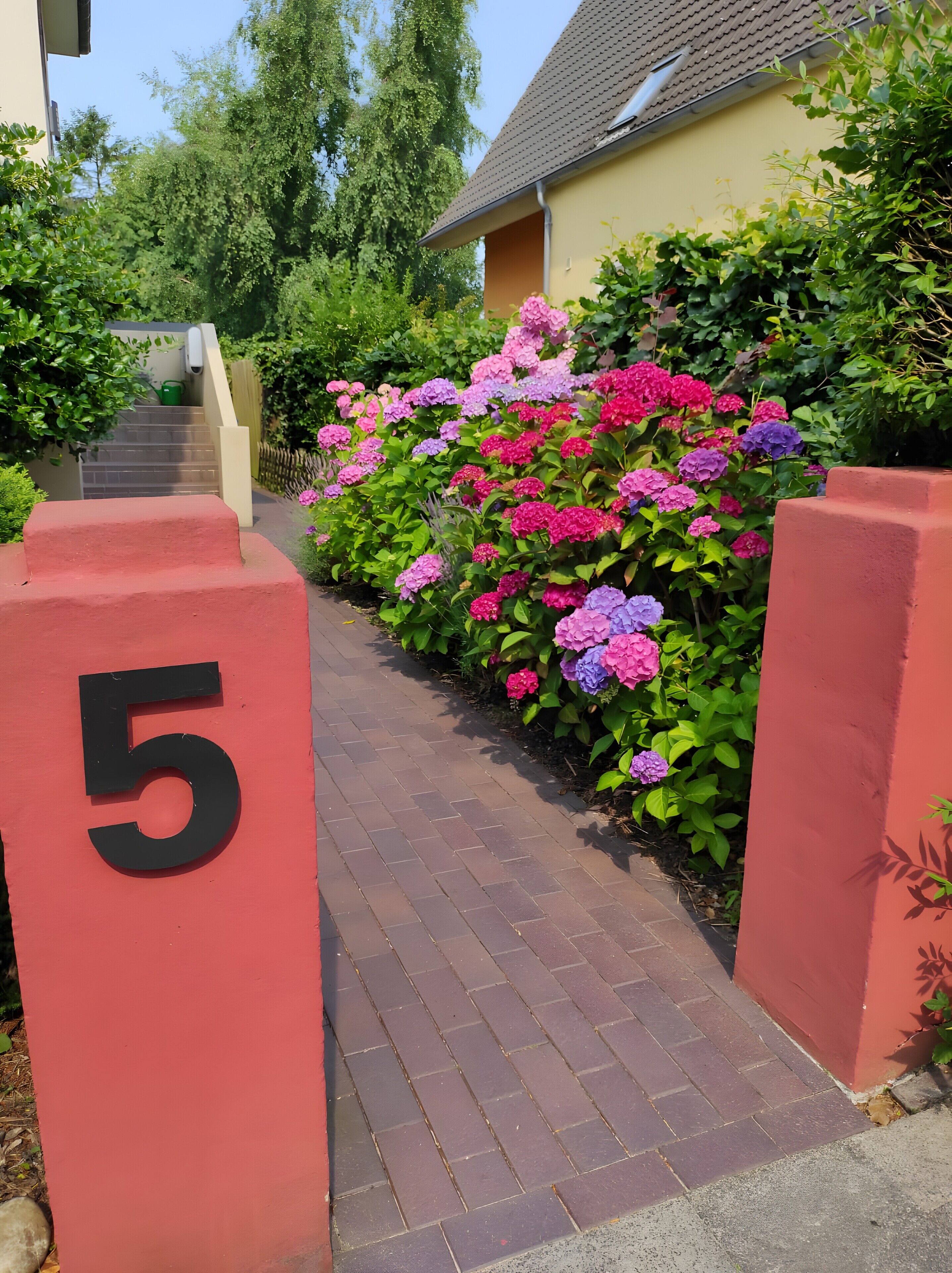 En stig leder fram till ett hus med nummer 5 på dörren, och huset är omgivet av frodig grönska och färgstarka blommor.
