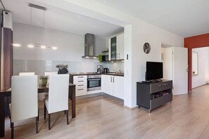 A modern kitchen with white cabinets, a microwave, and a stove. There is a dining area with a table and chairs. A TV is on a stand in the living area.