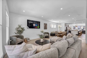 A modern living room with a large flat-screen TV, comfortable seating, a dining area, and a kitchen in the background.
