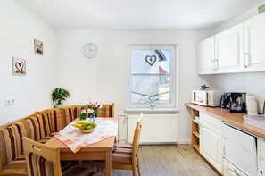 A compact kitchen with white cabinets, a dining table with a bench, a clock, and a window with a heart decoration.