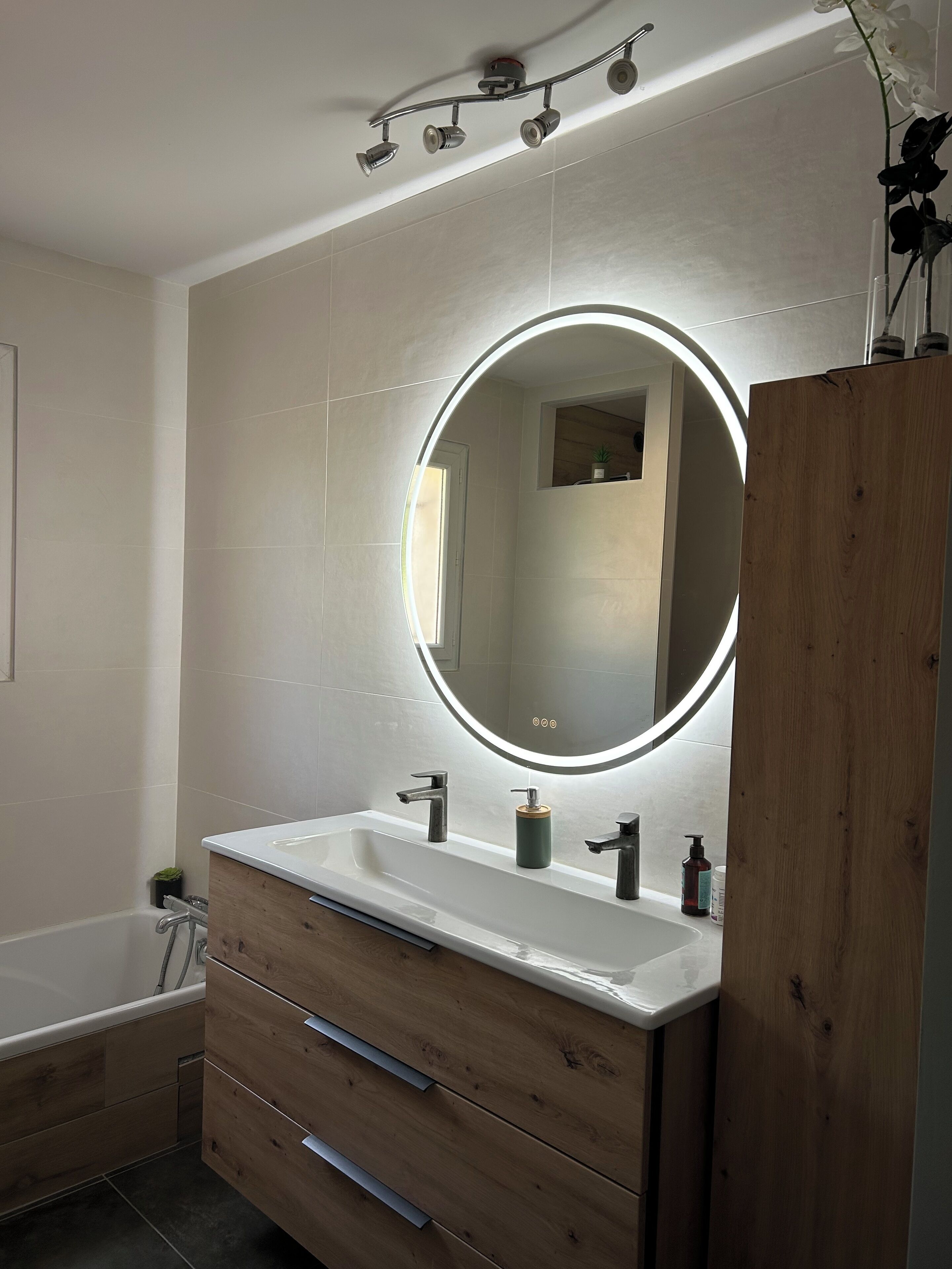 Un baño moderno con tocador de madera, un espejo redondo con luz y una bañera.