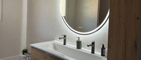 Un baño moderno con tocador de madera, un espejo redondo con luz y una bañera.