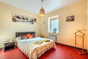 A bedroom with a bed, bedside tables, a hanging light, artwork, a window, and a radiator.