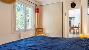 A bedroom with a bed, a chair, a window, a wardrobe, and a dining area visible through an open door. - Family House 400m from the Beach - La Grière Plage (La Tranche sur mer)
