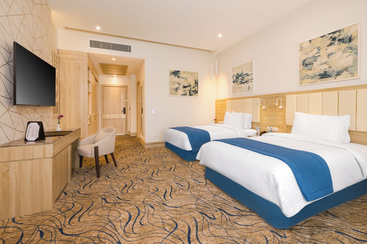 A hotel room with two beds, a desk, a chair, a flat-screen TV, and artwork on the walls.