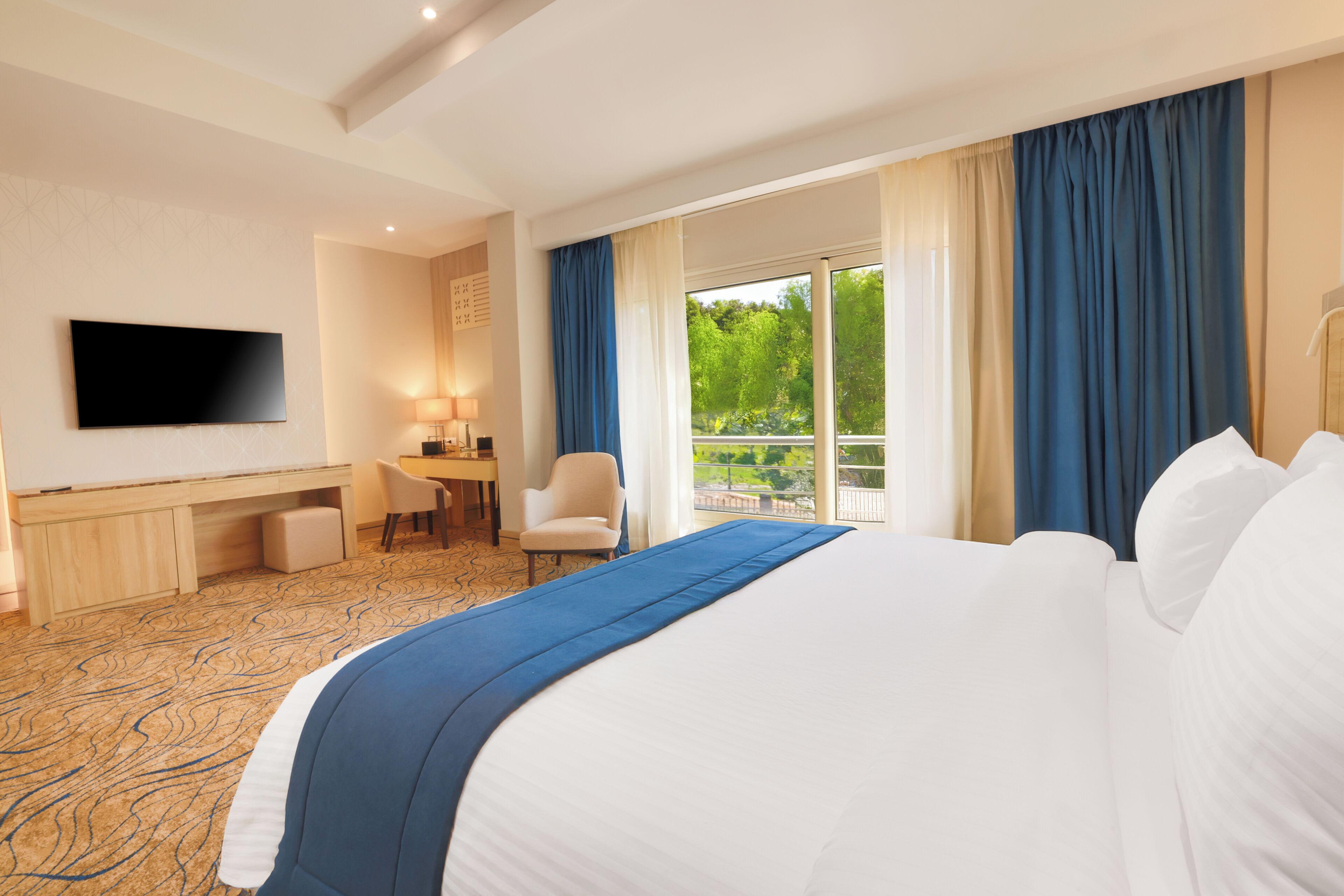 A hotel room with a large bed, a desk, a chair, a TV, and a balcony with curtains.