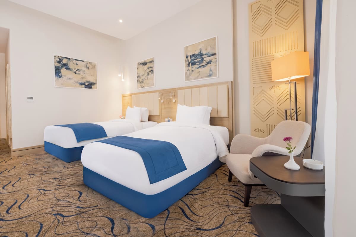 A hotel room with two beds, a chair, a small table with a vase, and abstract wall art.