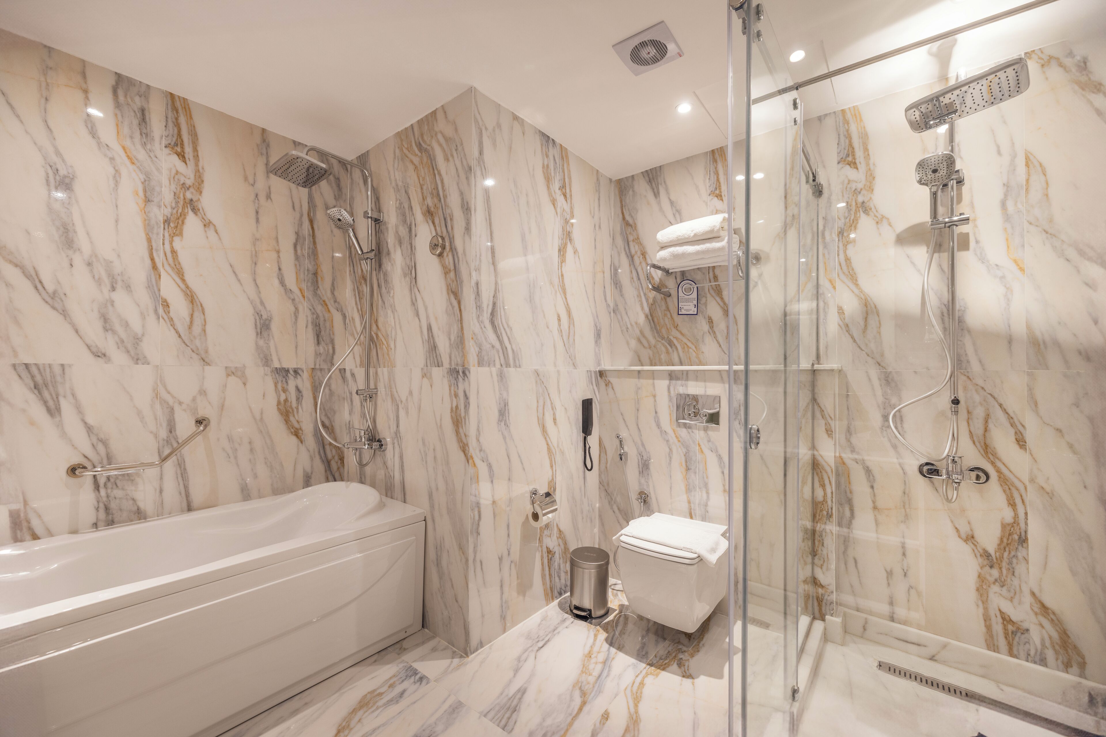 A modern bathroom with a marble wall, a bathtub, a toilet, and a glass-enclosed shower area.