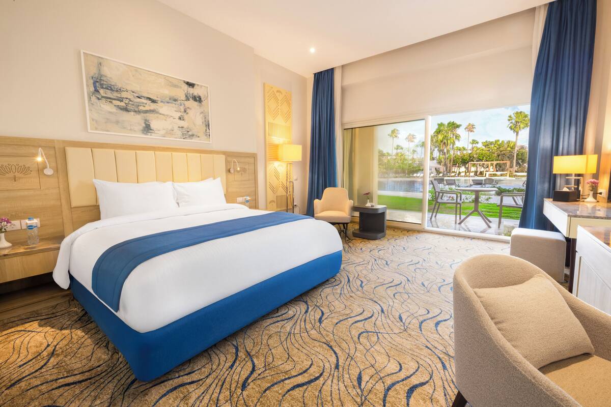 A hotel room with a large bed, a desk, a chair, a view of the pool, and a balcony with curtains.