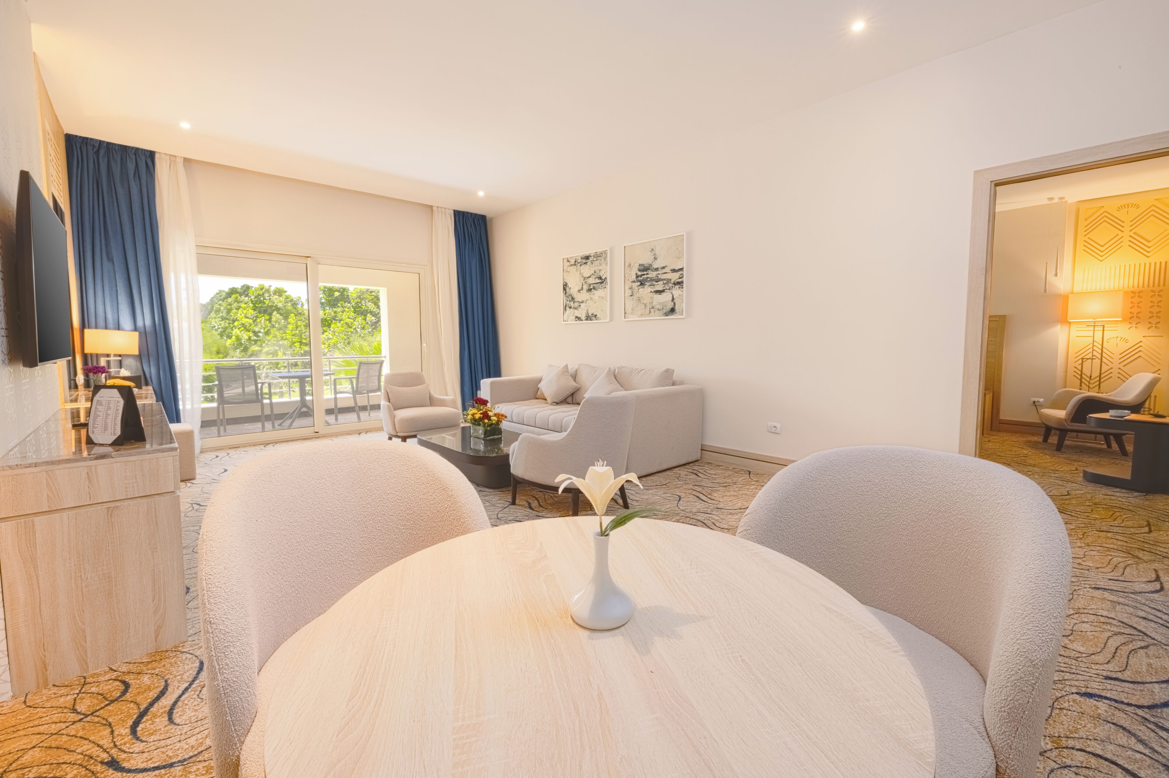 A modern hotel room with a sofa, a round table with a vase, a TV, and a balcony with a view of greenery.