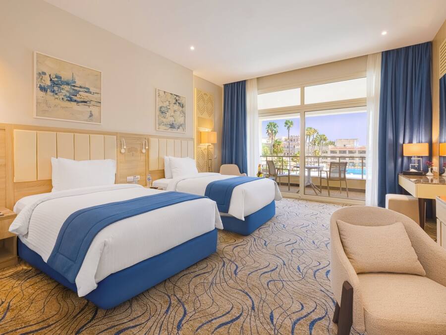 A hotel room with two beds, a sofa, a desk, and a balcony with a view of palm trees and buildings.