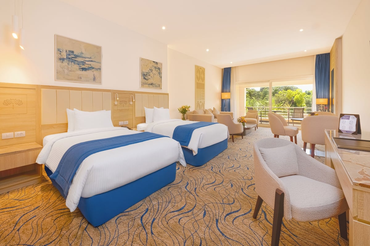 A hotel room with two beds, a desk, chairs, and a large window with a view of greenery.