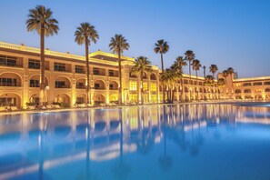 Un hôtel doté d’une grande piscine, de palmiers et d’un style architectural classique.
