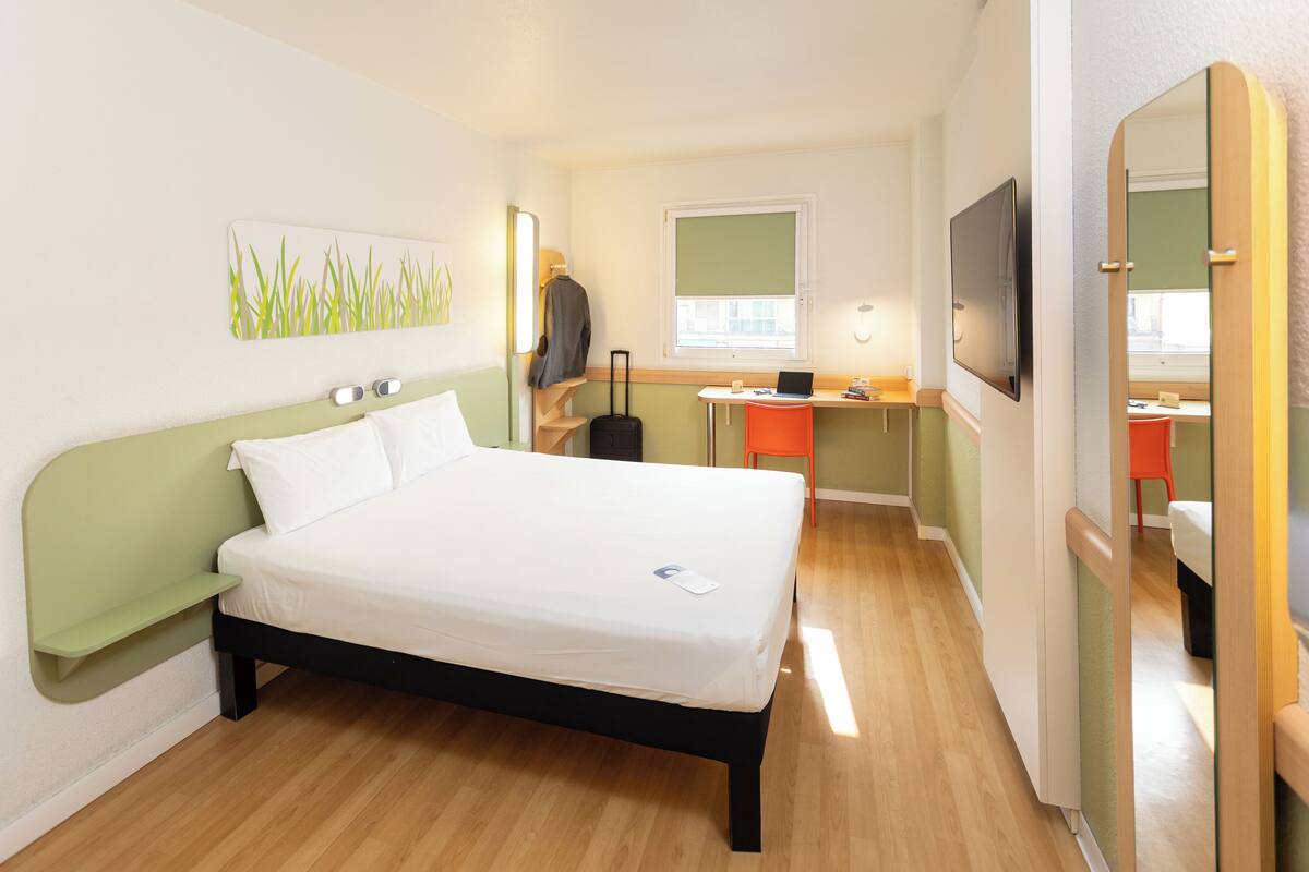Une chambre d’hôtel avec un lit, un bureau, une chaise, une télévision et une valise.