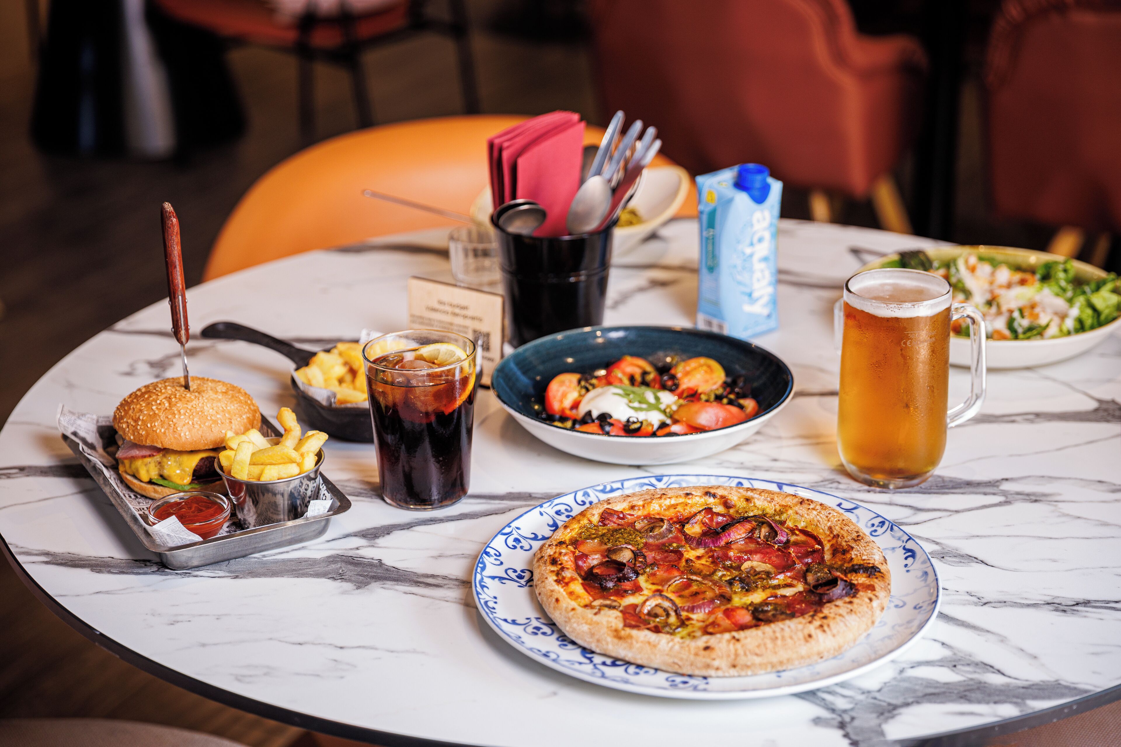 Una mesa de comedor con una hamburguesa, papas fritas, un vaso de refresco, una jarra de cerveza, una pizza y una ensalada.
