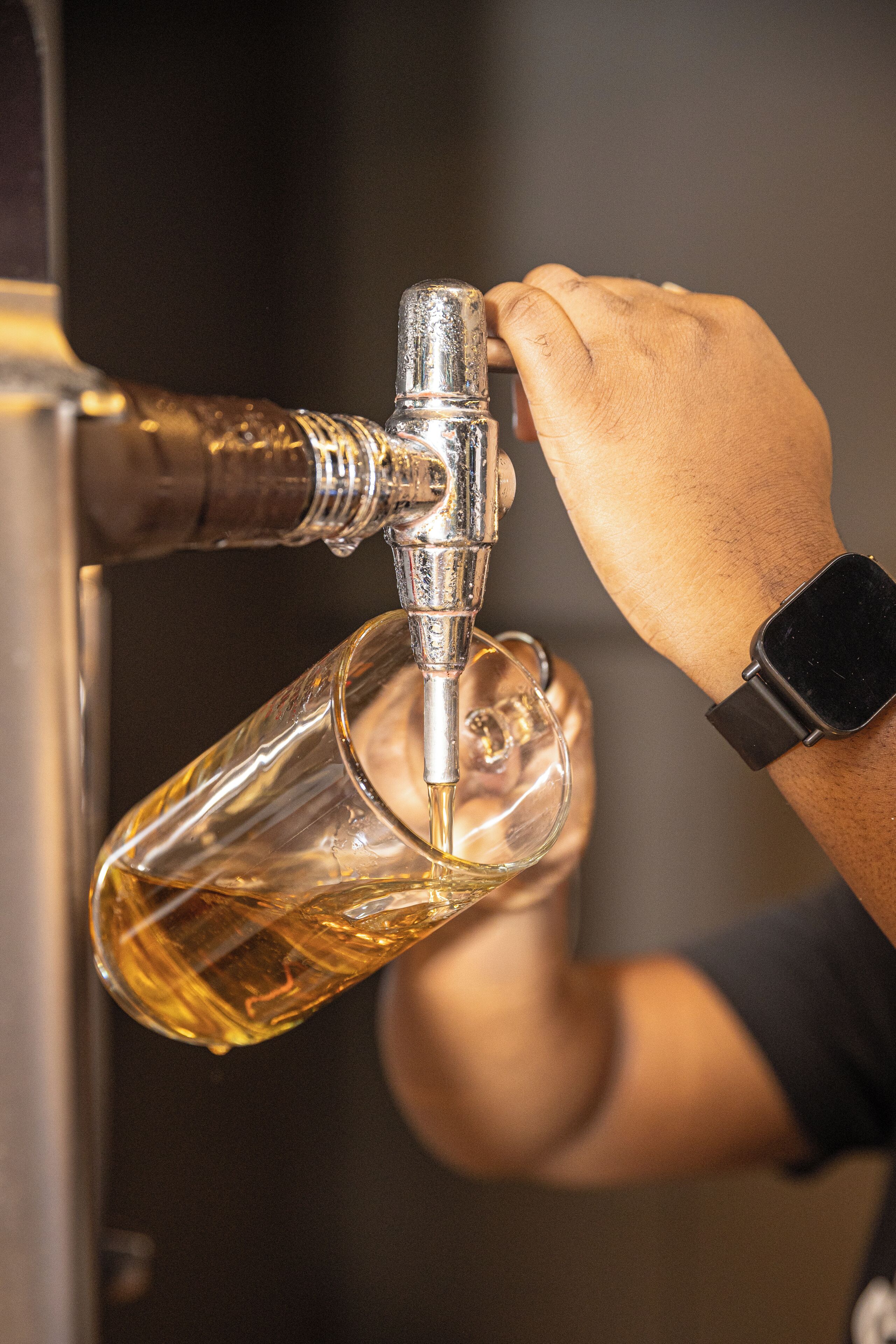 A hand pouring a golden liquid from a tap into a clear glass.