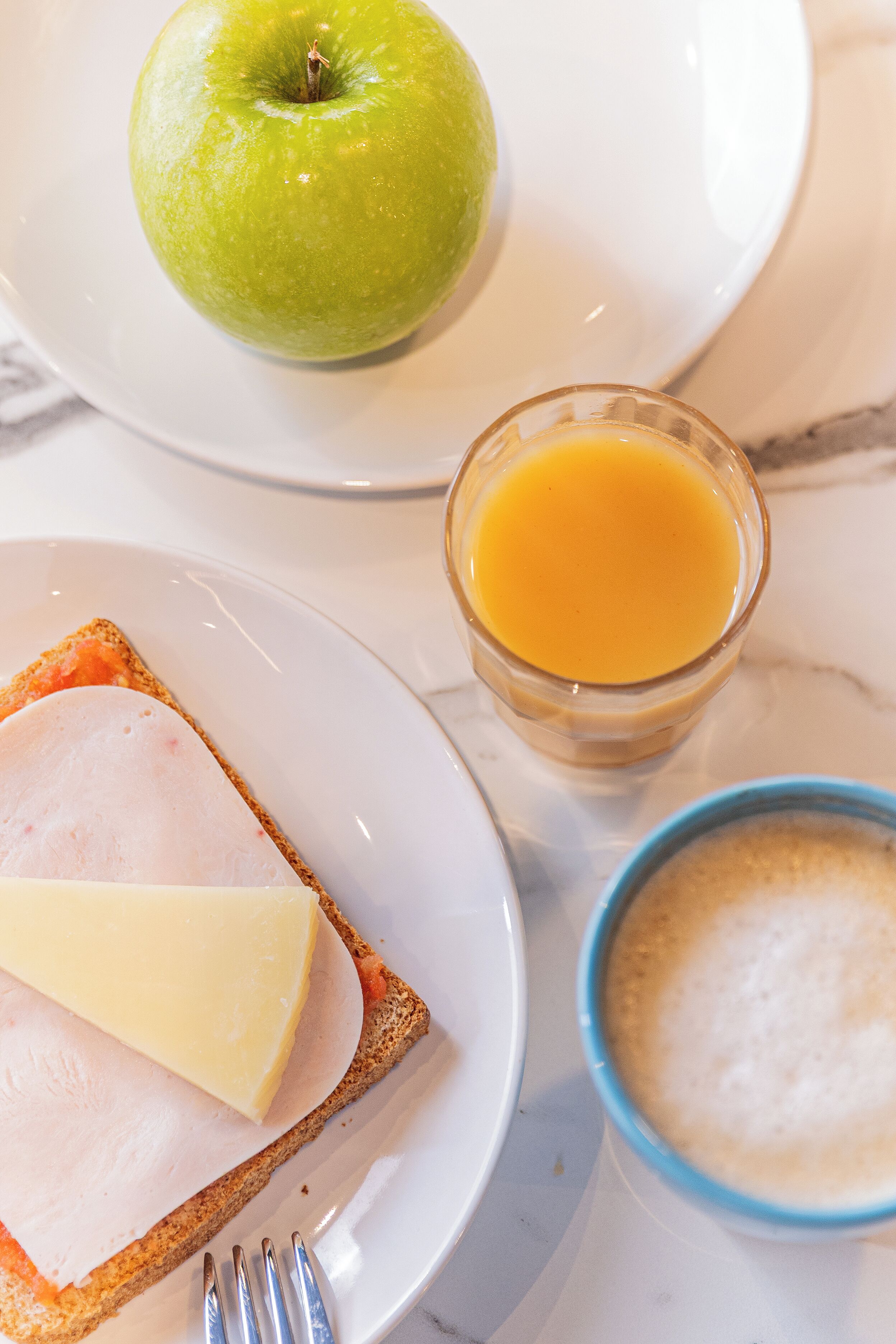 Ein Frühstück mit Toast, Schinken, Käse, einem Apfel, einem Glas Orangensaft und einer Schüssel Müsli.