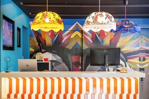 A reception desk with a laptop, a monitor, and a sign. There are two hanging lamps with floral patterns. The wall behind has a colorful geometric design.