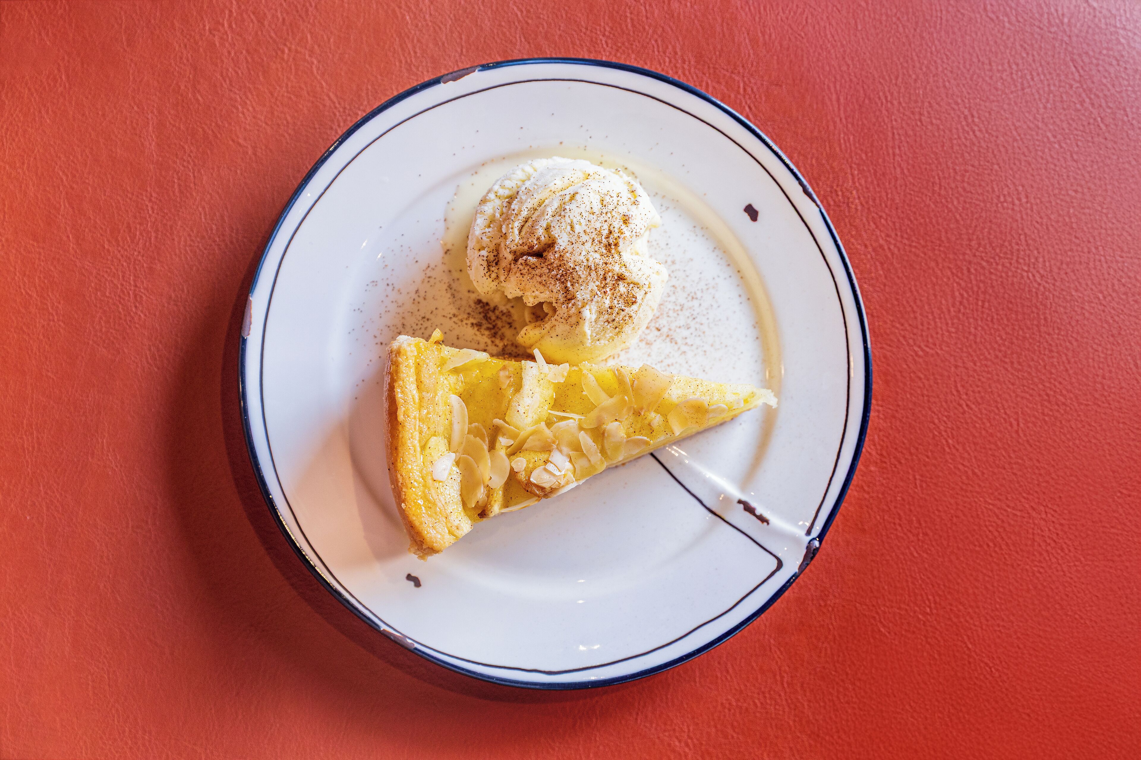 Un plato con una porción de pastel y una bola de helado sobre un plato blanco con borde azul, sobre un fondo rojo.
