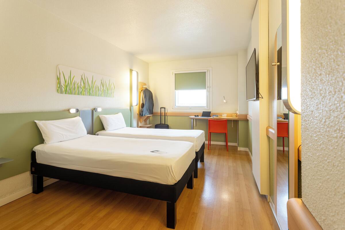 Une chambre d’hôtel avec deux lits, un bureau, une chaise, une télévision et une fenêtre.