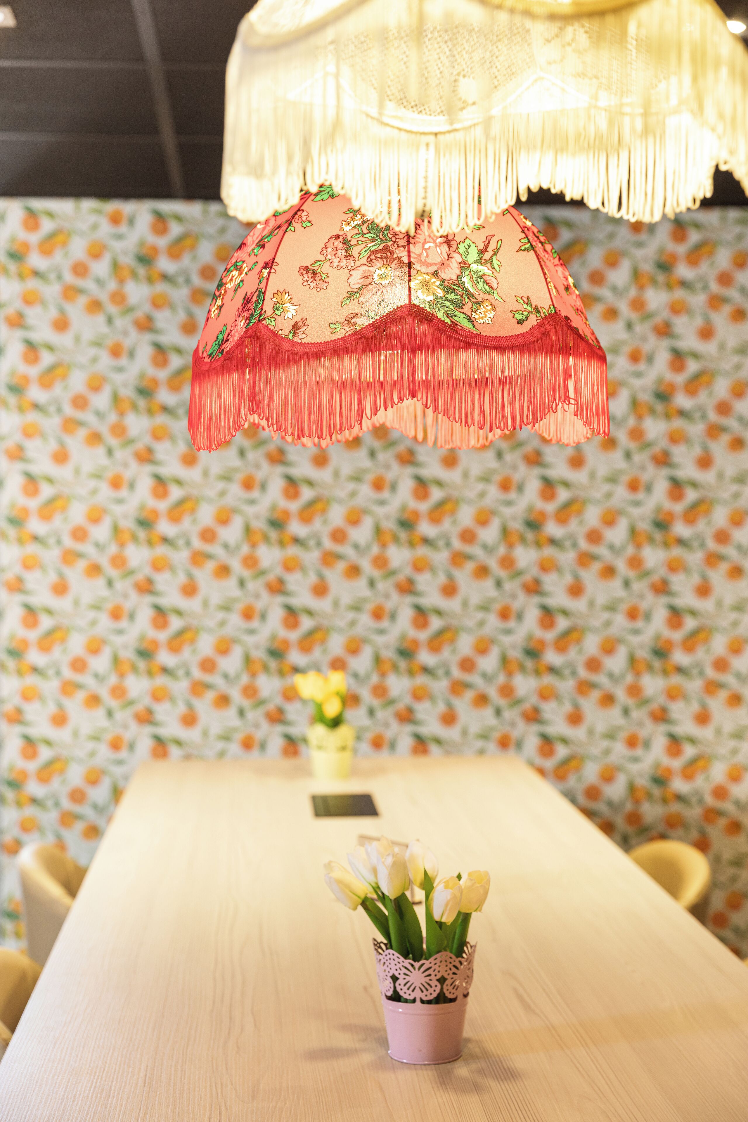 A table with a vase of yellow tulips, a hanging red fringe umbrella, and a patterned wallpaper background.