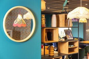 A room with a teal wall, a round mirror with a floral lampshade, and a wooden bookshelf with various items.