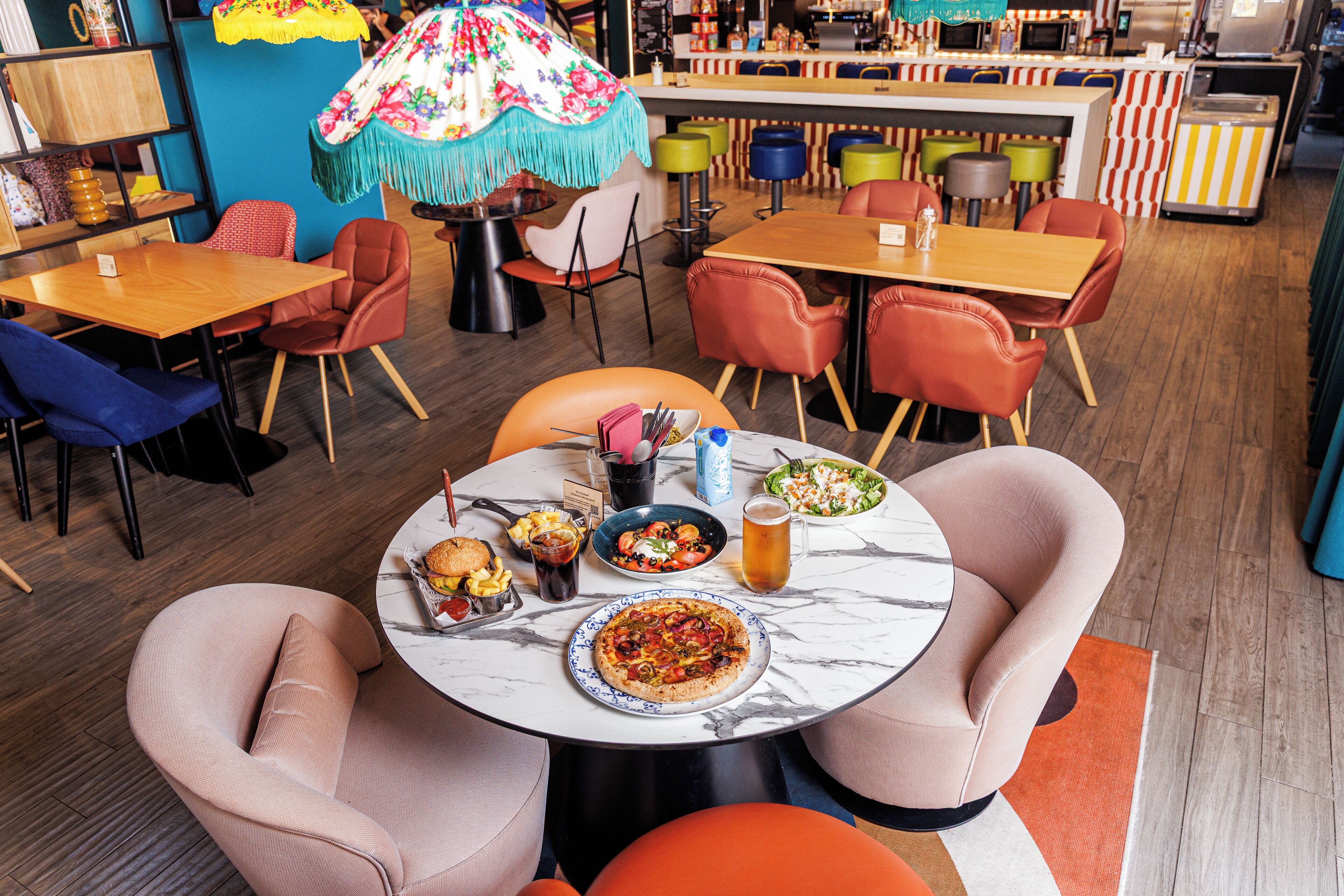 A restaurant with a marble table, pizza, salad, and drinks. There are colorful chairs and a bar in the background.