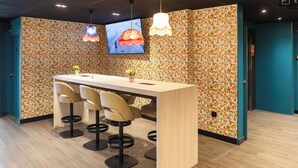 A modern bar area with a wooden counter, four bar stools, and a TV screen displaying a person running. - Ibis Budget Valencia Aeropuerto (Manises)