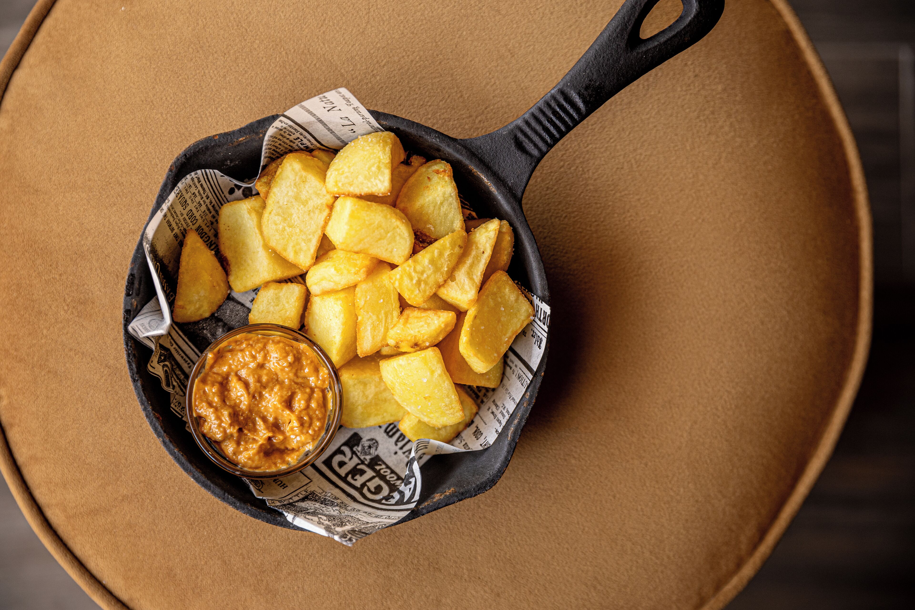 A cast iron skillet filled with golden potato wedges and a bowl of dipping sauce, placed on a brown surface.