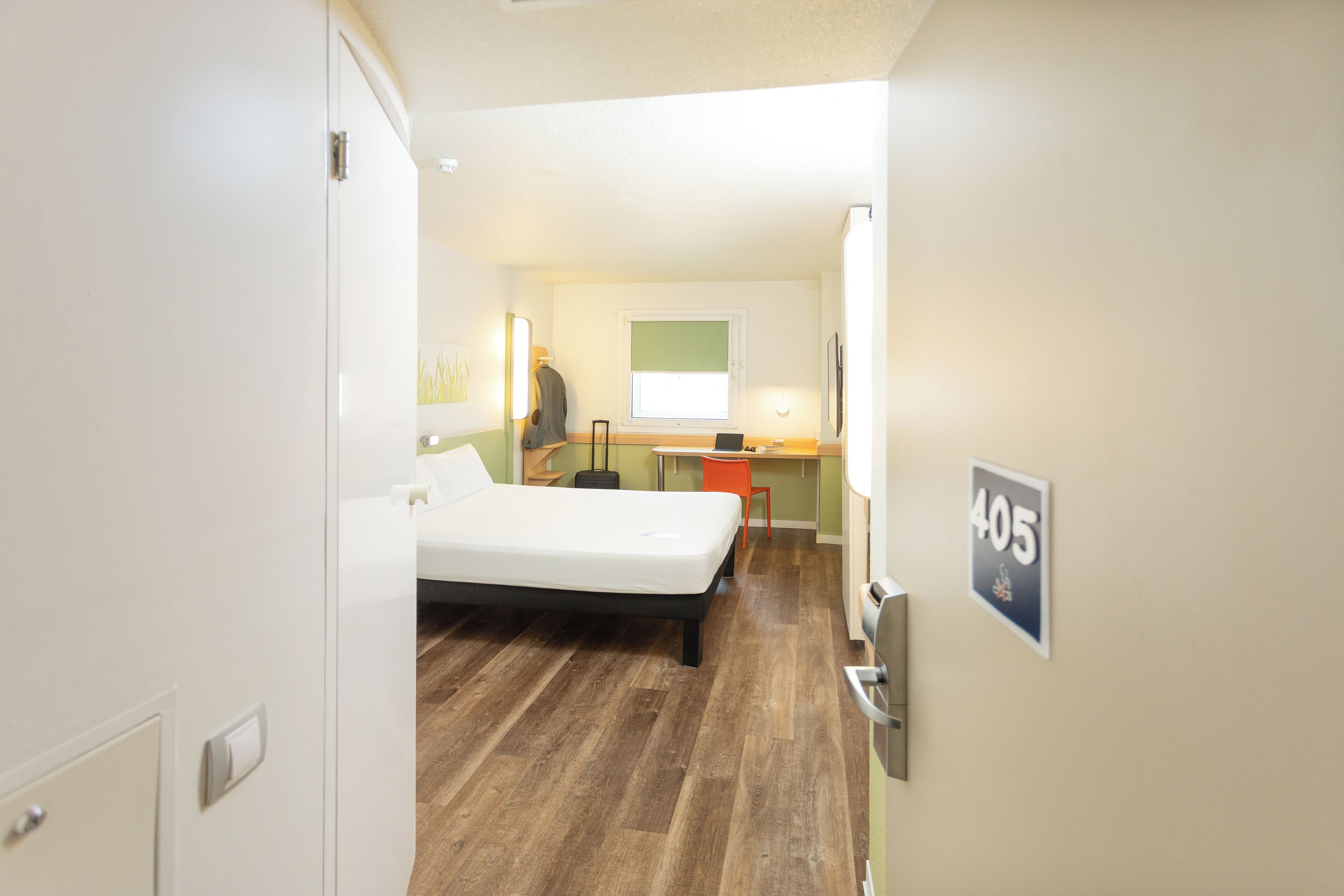 Habitación de hotel pequeña con una cama individual, un escritorio y una silla. Se ve una ventana con persianas, y el número de habitación 405 está indicado en la pared.
