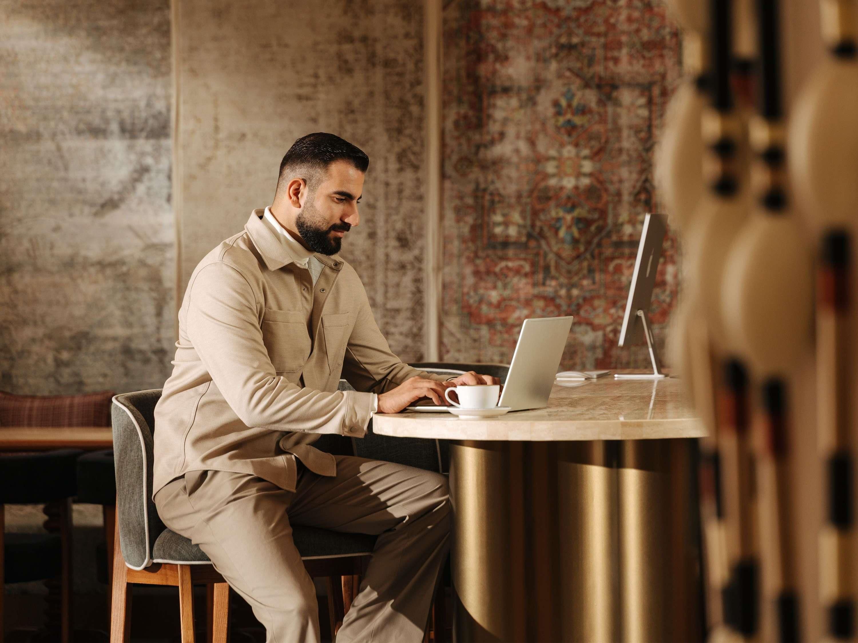 Une personne est assise à une table ronde, un ordinateur portable devant elle, dans une pièce dont le mur est décoré d’un motif et qui contient un banc en bois.
