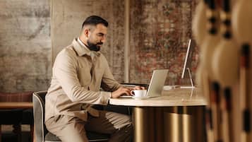 Une personne est assise à une table ronde, un ordinateur portable devant elle, dans une pièce dont le mur est décoré d’un motif et qui contient un banc en bois.