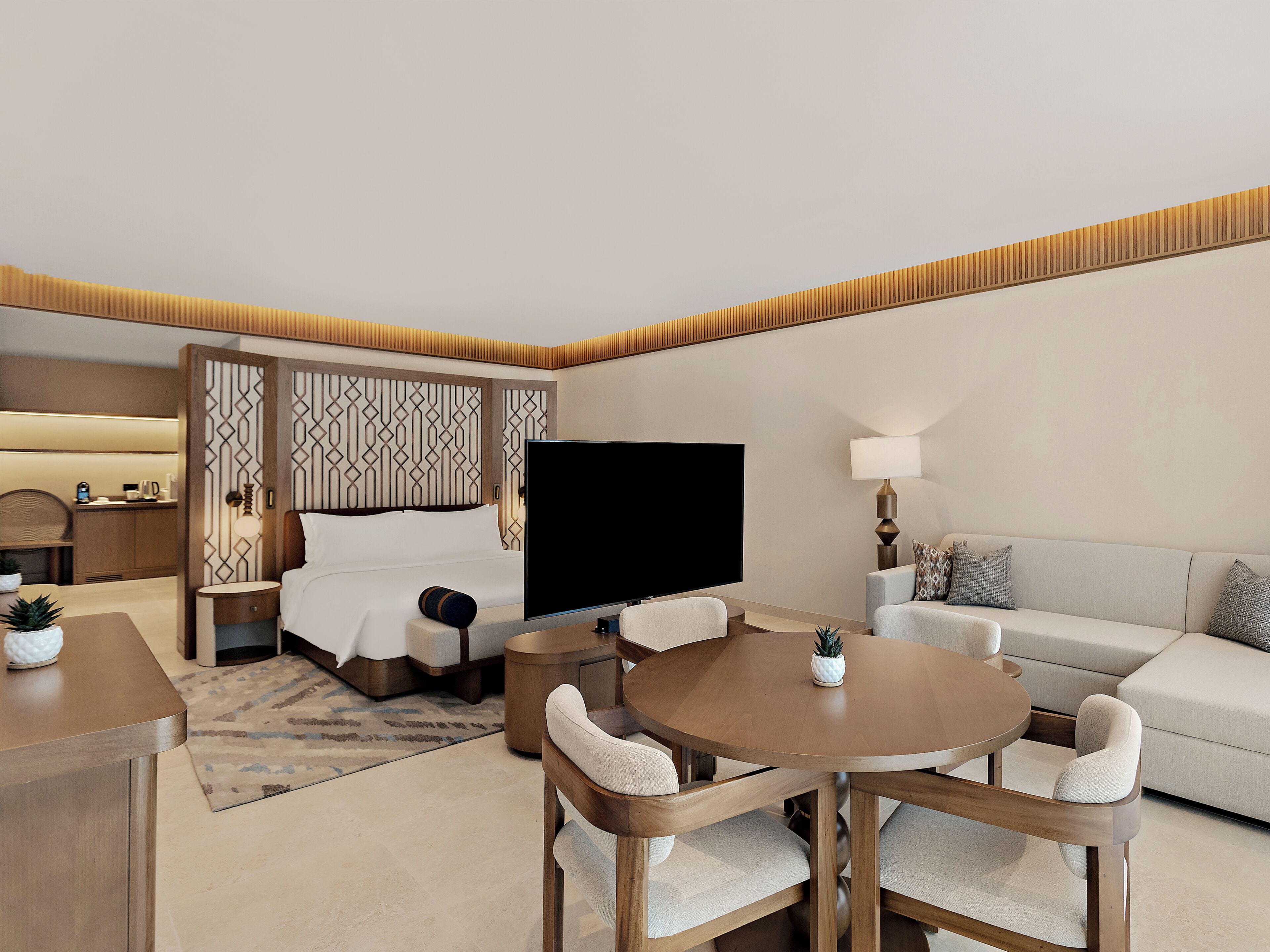 A hotel room with a bed, a TV, a sofa, a dining table with chairs, and a small table with a lamp.