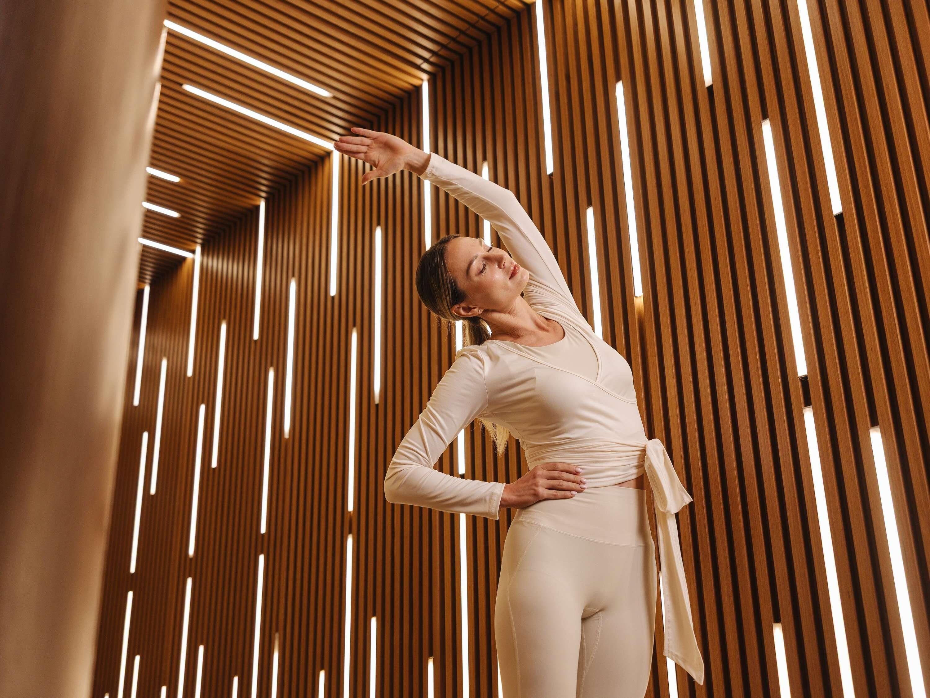 A person in a white outfit performing a stretching pose in a room with wooden slats and linear light fixtures.