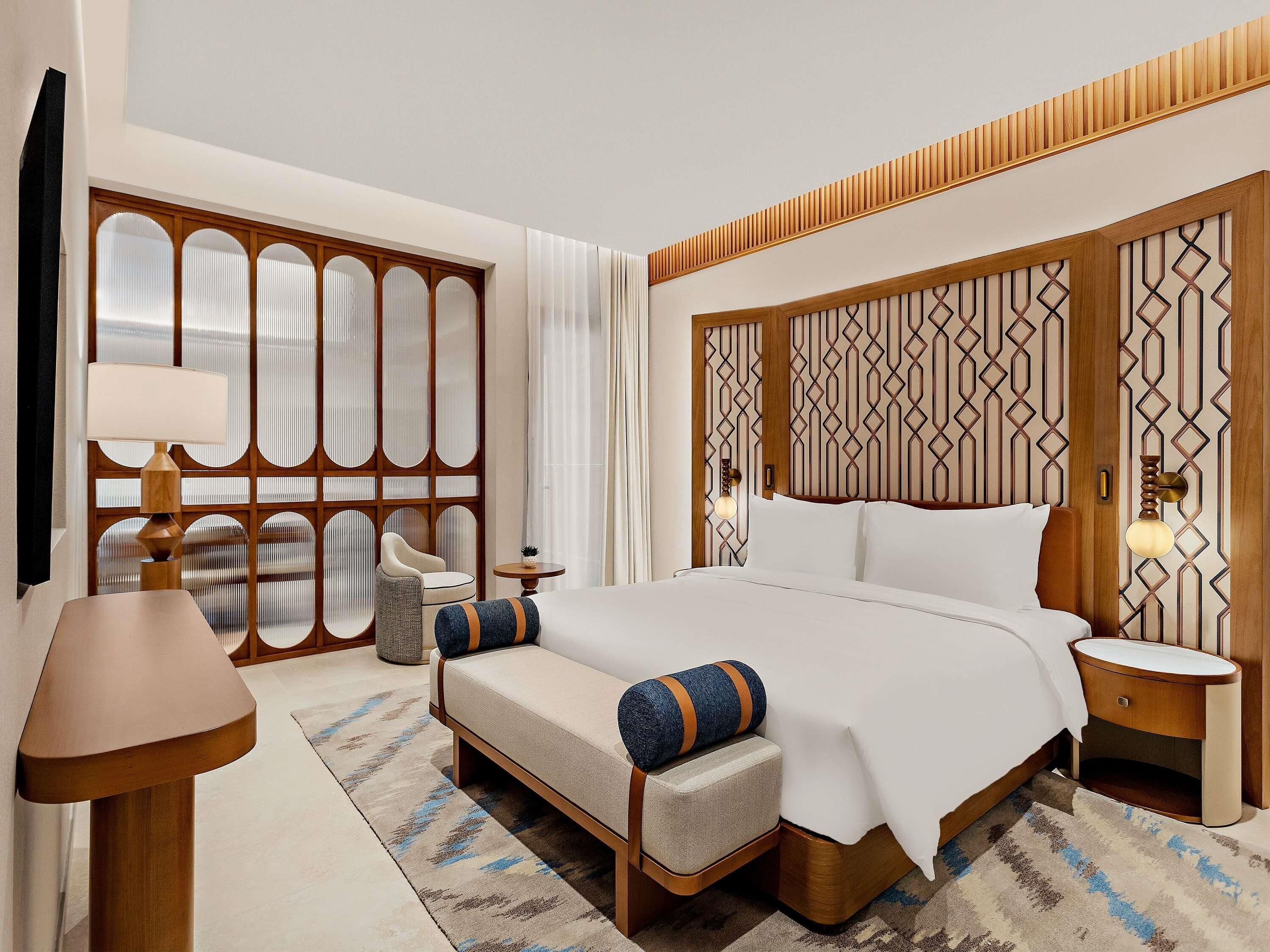 A hotel room with a large bed, a seating area with a chair and a small table, a lamp, and a decorative wall panel.