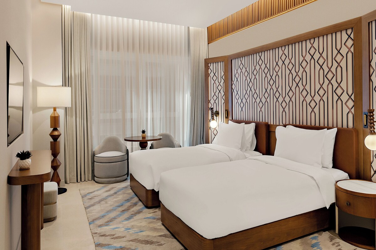 Habitación de hotel con una cama grande, dos mesitas de noche, una silla, una mesita pequeña y una lámpara.