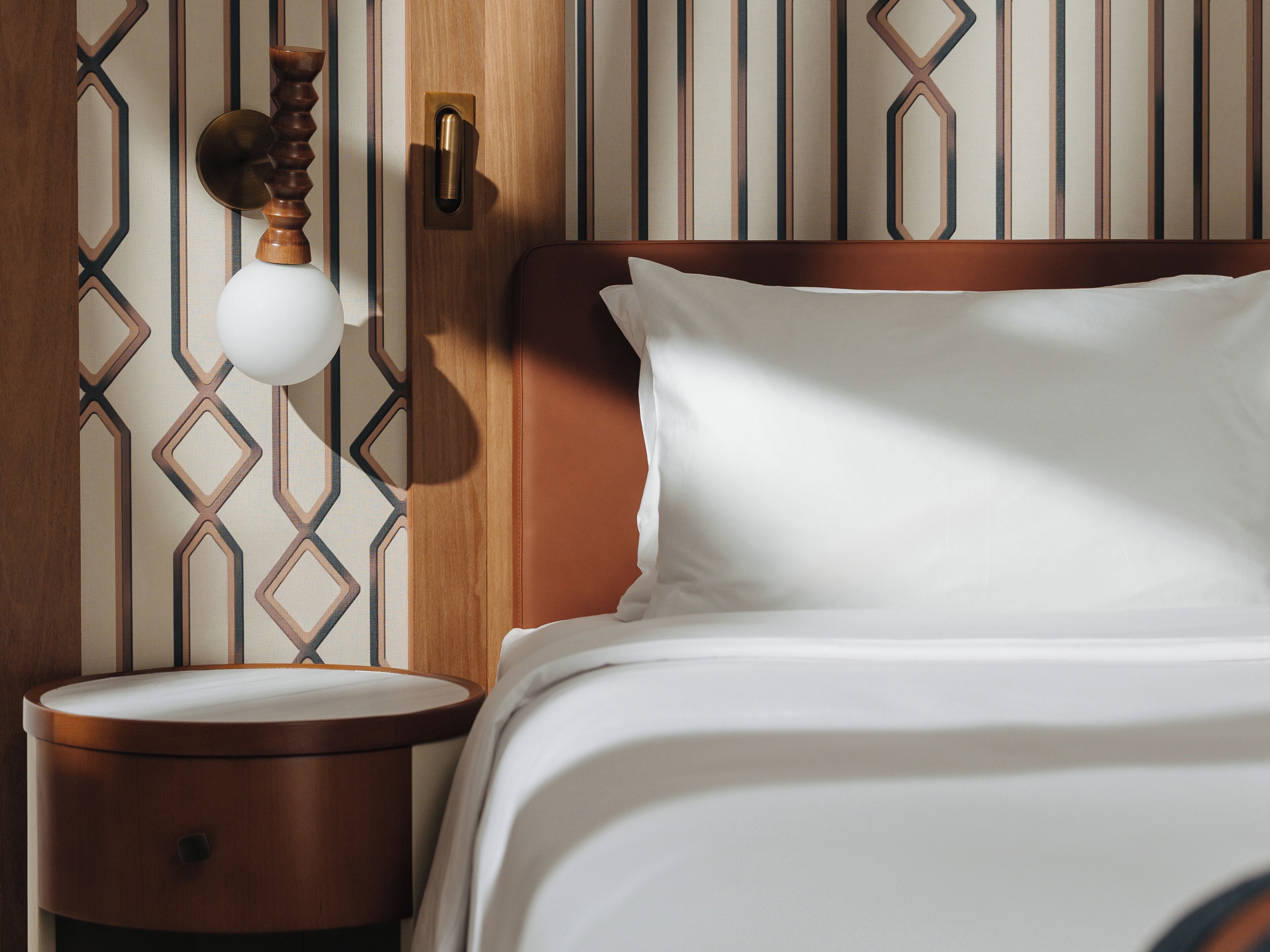 A neatly made bed with white linens, a wooden nightstand with a round drawer, and a wall with geometric patterned wallpaper.