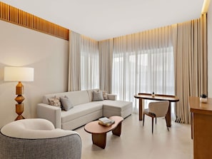 Een moderne hotelkamer met een bank, fauteuil, salontafel en een zithoek met tafel en stoelen.