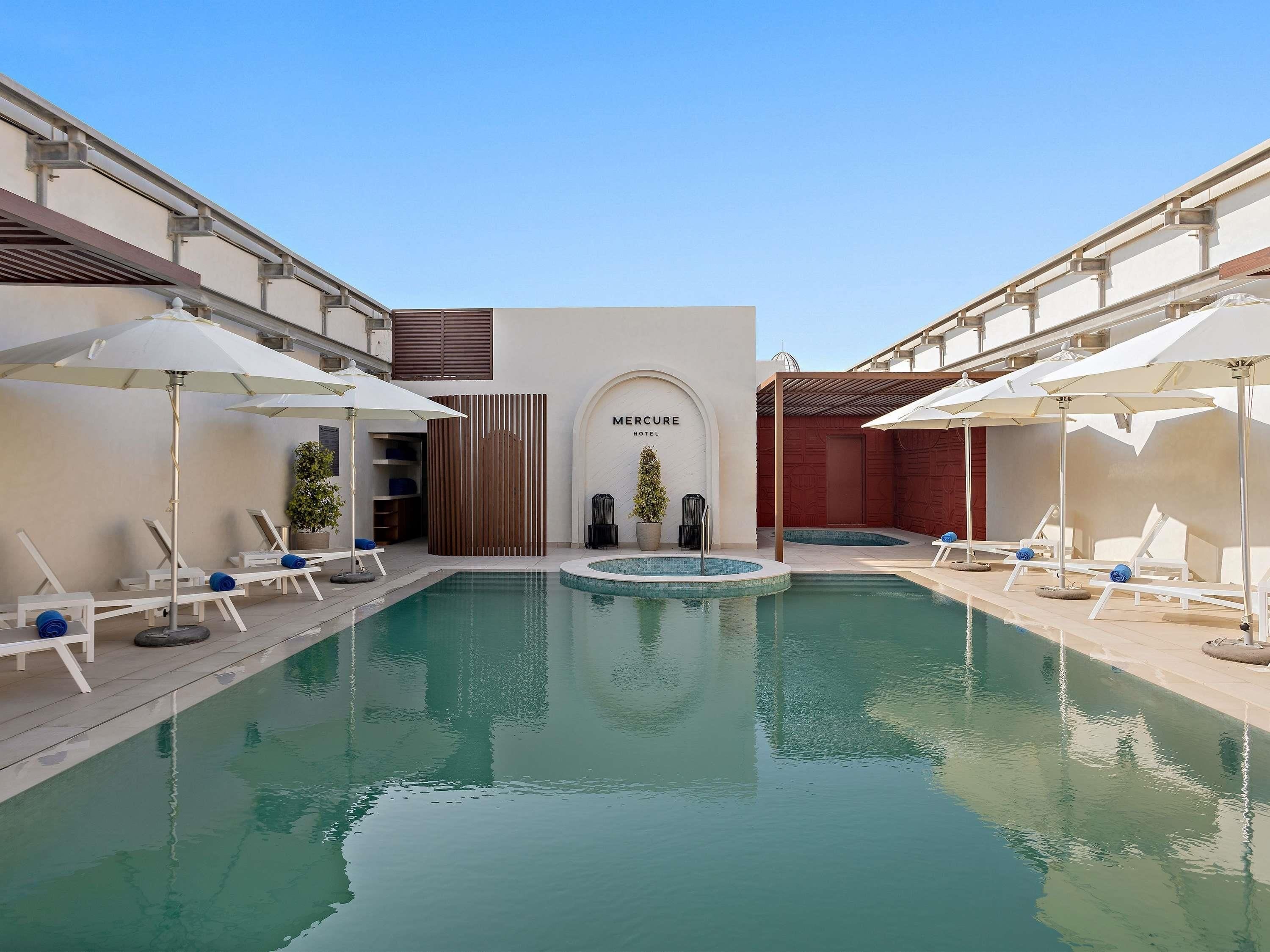 Un espace piscine avec des chaises longues et des parasols, un comptoir d’accueil et une enseigne indiquant « Hôtel Mercure ».