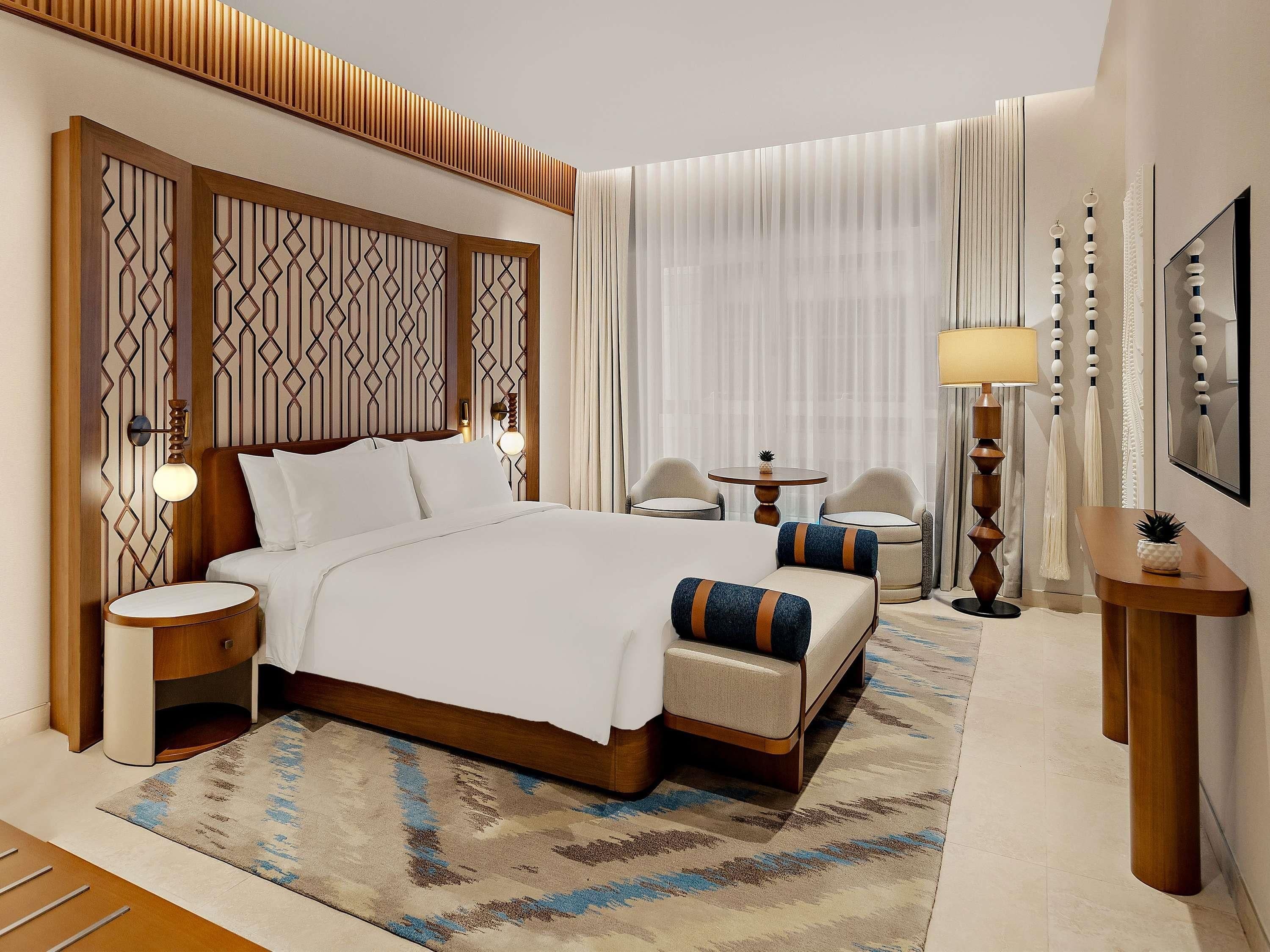 A hotel room with a large bed, two bedside tables, a small round table, two chairs, a floor lamp, and a decorative wall panel.