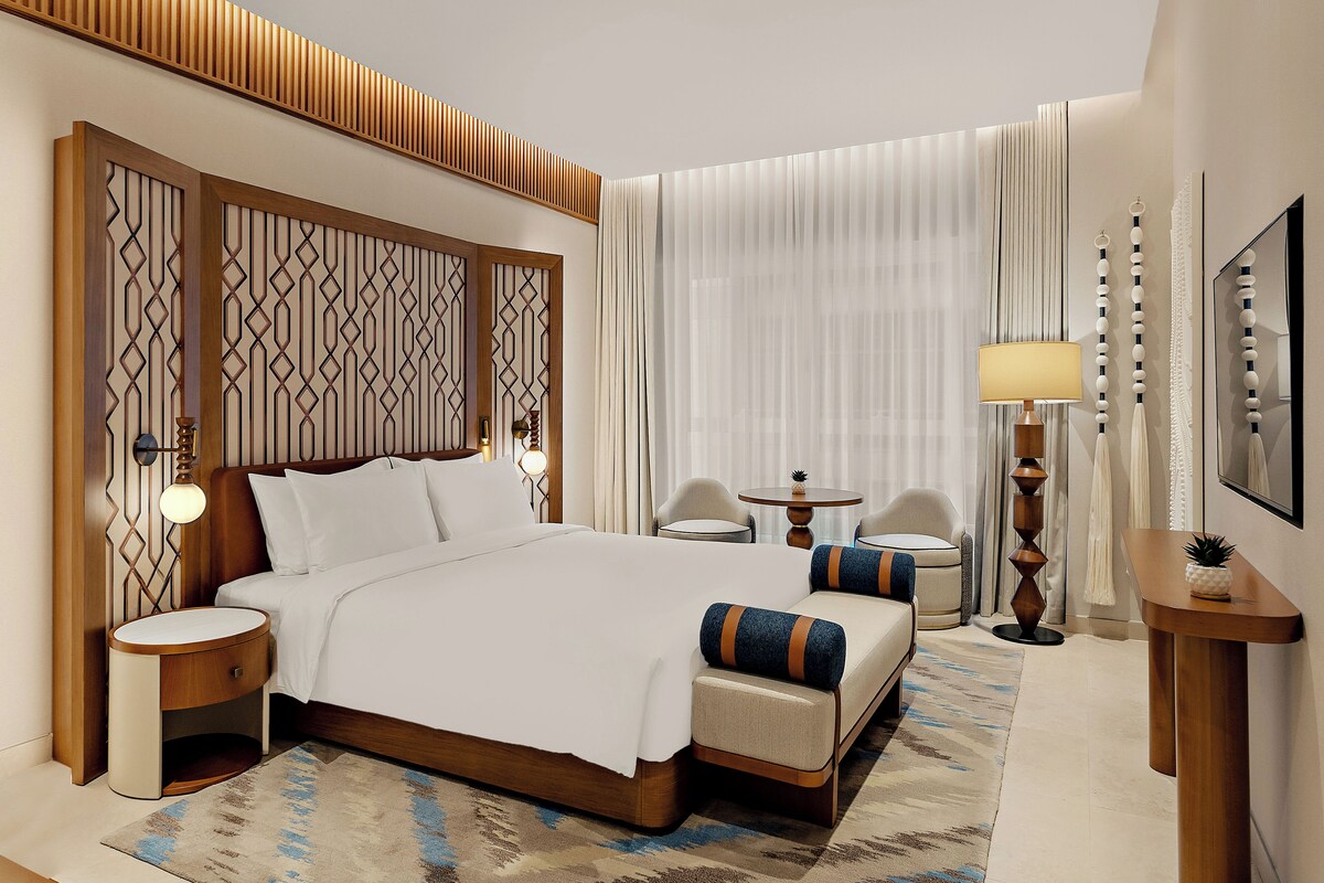 Habitación de hotel con una cama grande, dos sillas, una mesita, una lámpara y un panel decorativo en la pared.