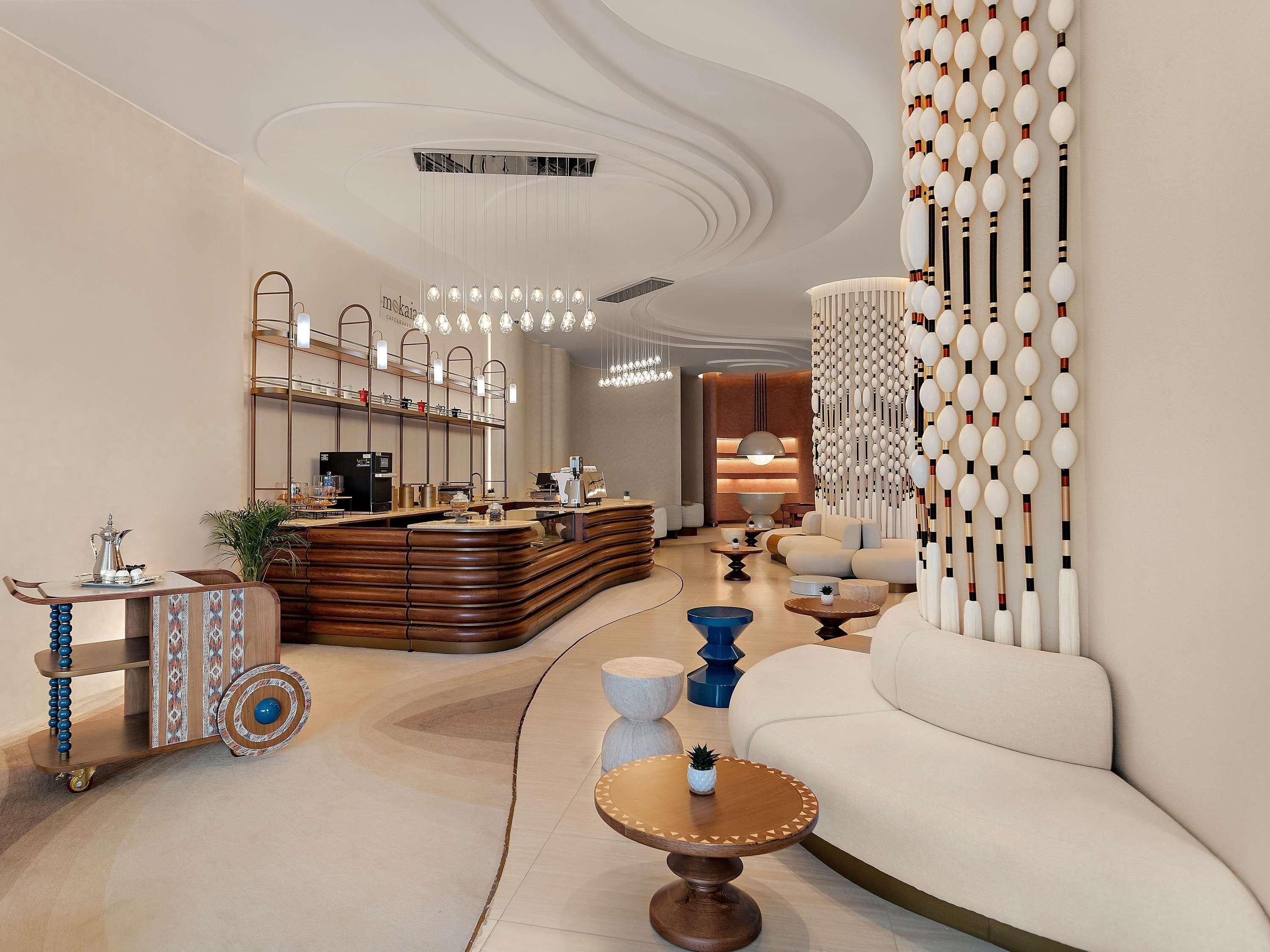 Um interior moderno com um balcão de recepção curvo, uma área de estar com um sofá e poltronas, e uma parede decorativa com elementos suspensos.